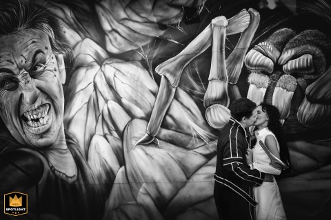 At the Saint Michel Festival in Toulouse, Haute-Garonne, the newlyweds share a kiss in cosplay attire, standing in front of a painted and illustrated amusement ride backdrop, captured in a black and white photograph that emphasizes their unique style.