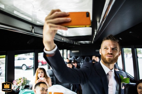 In San Francisco, California, the groom stands at the front of a party bus taking a selfie, while the group of seated guests behind him smile and pose, capturing a candid and energetic post-ceremony celebration.