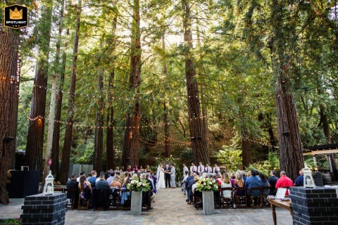 At Deer Park Villa in Fairfax, California, the bride and groom stand together at an outdoor altar beneath a towering canopy of trees, while guests are seated nearby, surrounded by the natural beauty and serene forest setting.