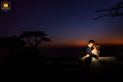In Carmel, California, the bride and groom pose for wedding portraits at sunset, dramatically illuminated by an off-camera flash used by the photographer, which adds depth and a romantic glow against the softly fading evening light.