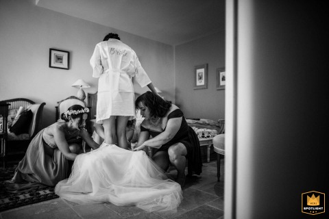 At Le Pré de l'Aube in France, the bride is gracefully stepping into her wedding dress, assisted by two attentive attendants, captured in a timeless black and white image that conveys anticipation and delicate care in the preparation suite.
