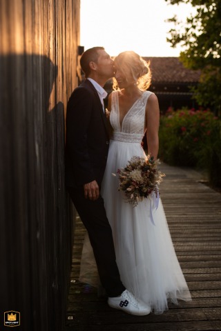 At Domaine de Beaulieu in Auch, the bride and groom stand close together on a boardwalk, illuminated by the sun behind them, sharing an intimate gaze against a rustic, wood-planked wall in the Gers countryside.