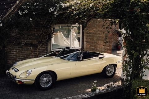Brides Reflection Appears Elegantly In The Window Of A Vintage Jag Wedding Car In Sussex UK In Sussex, UK, the bride’s image softly appears as she walks past the vintage Jag wedding car, her reflection elegantly captured in the window on her way to join the celebration.