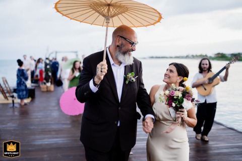 Couple Walks Along The Pier In The Rain Leaving Their Playa Del Carmen Ceremony In Mexico Leaving their Playa del Carmen ceremony, the couple walks along the pier as musicians and guests follow them in the rain, blending joy with the spirited energy of the celebration.