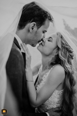 Soft light and a sheer veil envelop the bride and groom at a Budapest ceremony location as they lean in for a gentle kiss, their affection highlighted in an intimate setting.