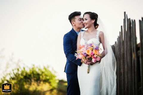 Coastal Portrait Of A Couple Before A Rustic Picket Fence Under Blue Skies In Capitola California A couple poses for a wedding day portrait in Capitola, CA, standing before a rustic wood picket fence under a vibrant blue sky, creating a picturesque, coastal celebration scene.