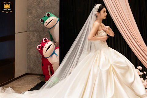A bride waits patiently in line at a hotel in Fujian, China, accompanied by two groomsmen playfully dressed as large frog mascots during a unique and humorous wedding moment.