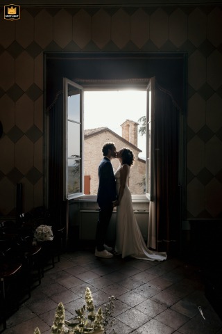 Inside the Longiano City Council in Italy, a couple shares a backlit silhouette kiss. Empty historic chambers and strong light from the windows give a cinematic impression to this quiet interlude.