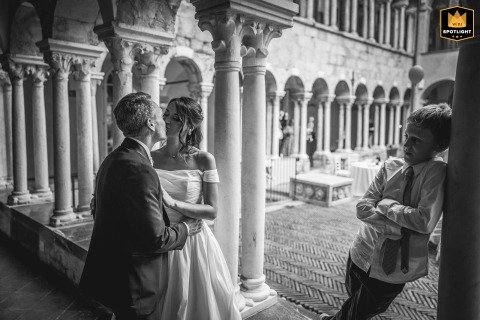In Genova, Italy, a black and white photo shows a couple kissing among rows of old stone columns. In the foreground, a boy stands with arms crossed, looking unimpressed and providing a humorous touch to the classic setting.