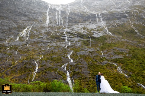 In Milford Sound within New Zealand’s Fjordland National Park, the newlywed couple walks hand in hand before a towering wall of waterfalls, their figures small against the dramatic backdrop of cliffs and cascading water.
