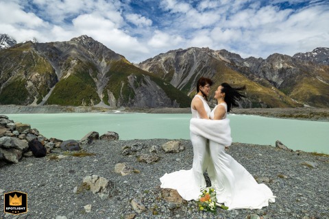 In Mt Aoraki National Park, New Zealand, two brides brace together against strong mountain winds, embracing to keep warm amid dramatic alpine scenery, their connection standing resilient against the power of the landscape.