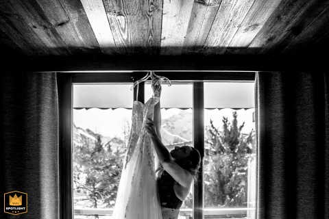 Alpine Window Scenery Contrasts Softly With A Focused Bride Picking Up Her Dress In Les Deux Alpes France In Les Deux Alpes, France, the bride reaches to pick up her wedding dress, the alpine scenery outside the window softly contrasting with her focused preparation inside the venue.