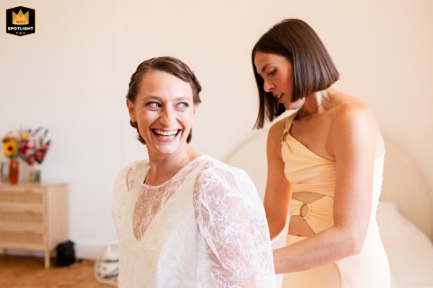 Soft Window Light Warms A Radiant Bride Getting Dressed At Domaine Du Mey Gers France At Domaine du Mey in Gers, France, the bride smiles radiantly during her wedding preparations as a bridesmaid fastens the back of her dress. Soft light from nearby windows adds warmth and highlights her excitement as she prepares for the ceremony.