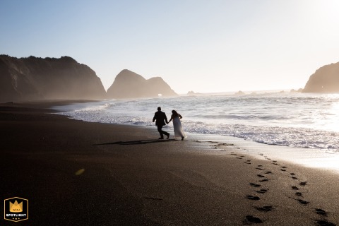 The Newlywed Couple Dashes Across The Windy Sand At Sunset Outside Elk Cove Inn Mendocino As sunset glows at Elk Cove Inn in Mendocino, the bride and groom dash across the sand in the wind.