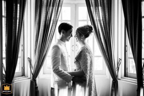 A black-and-white portrait at Baan Dusit Thani in Bangkok, Thailand, shows the couple standing symmetrically before windows with drawn-back curtains, creating a refined image defined by balance, light, and architectural elegance.