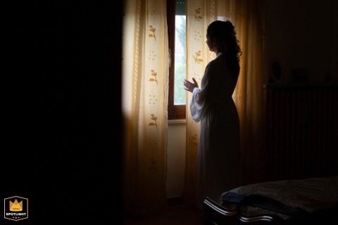 During preparations at the bride’s home in Brusatasso, Mantova, Italy, the bride gently parts a curtain to check the stormy weather conditions outside before the ceremony.