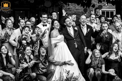 The bride and groom pose outdoors with a large group of guests at Mariënhof Amersfoort, as friends and family raise their hands and give thumbs-up gestures in celebration.