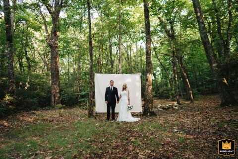 In the Blue Ridge Mountains of North Carolina, a bride and groom stand hand in hand before a white fabric backdrop, blending rustic forest calm with editorial portrait styling.