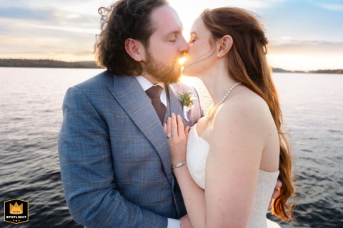 In Oquossoc, Maine, the bride and groom stand at sunset on a lake, leaning toward a kiss as the water and sky glow around them.
