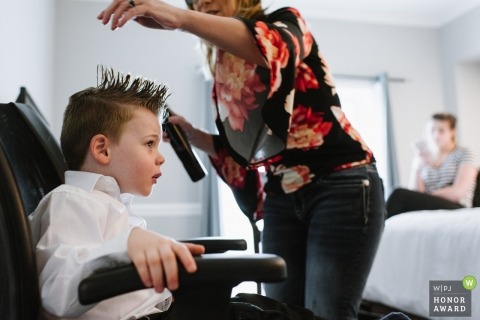 A small boy gets his hair spiked on top in a hotel room. The hair dresser leans over him while he sits in a hotel conference chair. 