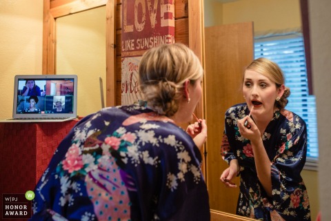Northern California wedding image of a bride as she puts on lipstick in her robe as her friends watch on via a Zoom call on a laptop
