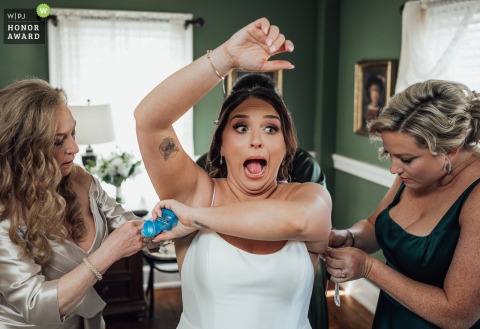 A bride at the Smithville Inn in Galloway, NJ, prepares for her wedding day with assistance in putting on her dress and applying deodorant. The moment was captured by a wedding photographer and received a WPJA Honor Award.