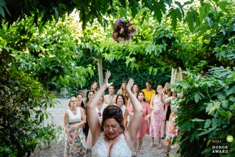 A skilled wedding photojournalist captured this award-winning image of a bouquet toss at Terres de Saint-Hilaire in France. The scene is surrounded by beautiful green foliage, creating a stunning frame.