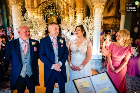 Bride bursts into laughter during her wedding ceremony at Wotton House, Surrey. The photograph captures her joy as the rest of the wedding party eagerly waits, curious about the source of her amusement. The image received an Honor Award from the WPJA.