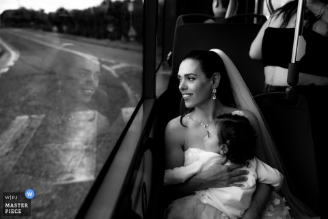 Forlì Bride's Reflective Transit to Ceremony with Child in Her Arms In Forlì, Italy, a bride travels to her ceremony on public transport, her thoughtful face reflected in the window, while holding a child.