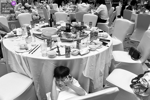 A young boy hides playfully under the wedding dinner table at the venue, captured in a candid, black and white moment.
