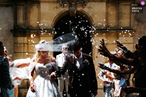 Bride with flying veil and groom exit the Château Coty, Tarn et Garonne, showered in petals, captured candidly in a documentary wedding moment.