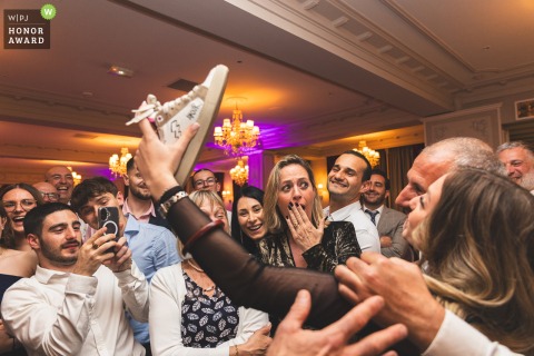 A hypnotized wedding guest at Hotel Servella Castagniers in France hilariously holds her shoe like a phone to take a selfie with a celebrity, captivating and amusing other guests.