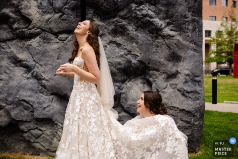 A bridesmaid crouches under the bride’s train, fixing her dress, creating a funny, candid moment at Denver Art Museum.