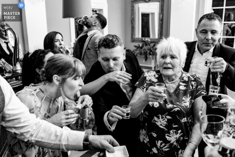 Wedding guests at Rushton Hall, UK, smiling and laughing while taking celebratory shots together at the bar during reception.