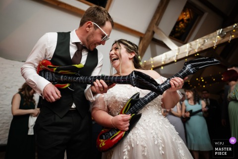 Bride and groom enthusiastically rock out with inflatable guitars, laughing and celebrating at Stratton Court Barn wedding reception.