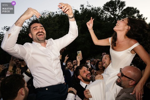 At Vandelli Garden, the bride and groom are joyfully lifted into the air by guests during a lively cocktail hour celebration.