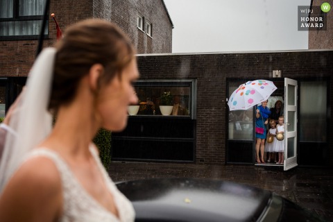   On a rainy wedding day outside the couple’s house, the bride climbs into the car while family members watch under umbrellas.