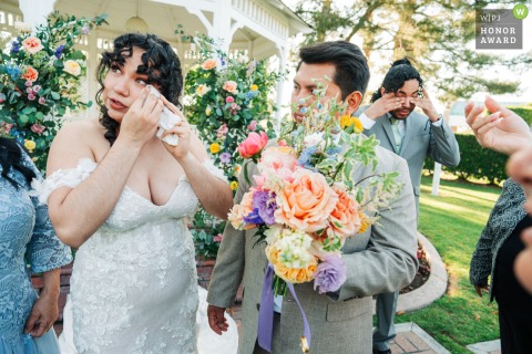   The bride wipes away tears while her groom holds her bouquet and her brother also gets emotional in the background at Alta Vista Country Club in Placentia, California.