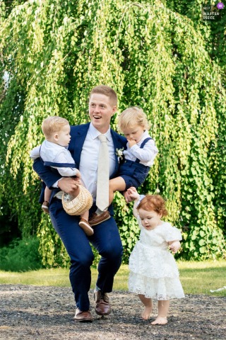 Groomsman Wrangles Flower Girls and Ring Bearers Down the Aisle in Wiscasset, Maine Wedding In Wiscasset, Maine, a groomsman struggles to wrangle all the flower girls and ring bearers down the aisle. The award-winning photograph highlights the photographer's skill at documenting chaotic, and humorous moments.
