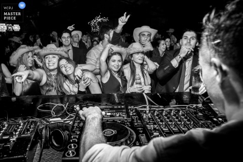 Rowdy Guests Dance in Cowboy Hats - DJ View at Rosario, Santa Fe Wedding In Rosario, Santa Fe, a black-and-white photograph, taken from over the DJ's shoulder, captures a rowdy crowd of guests dancing with party cowboy hats. The award-winning photograph highlights the photographer's skill at documenting energy.