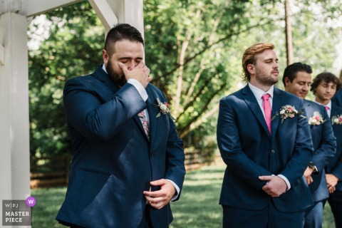 Groom Sees Bride for First Time Walking Down Outside Aisle at The Venues at Langtree, Mooresville At The Venues at Langtree in Mooresville, North Carolina, a groom sees his bride for the first time walking down the aisle. The award-winning photograph highlights the photographer's skill at documenting authentic emotions.