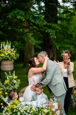 At Mulino dell'Olio in Clivio, Italy, newlyweds share a kiss after exchanging rings, with their children and an applauding celebrant in the background. The award-winning photo highlights the photographer's skills.
