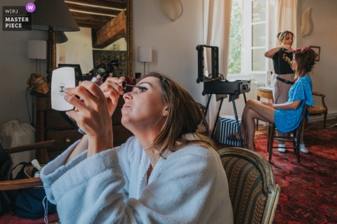 Bride Applies Makeup With Handheld Mirror While Another Gets Hair Styled At Maisonnais-sur-Tardoire Gironde At a home in Maisonnais-sur-Tardoire, Gironde, a bride applies her own makeup with a handheld mirror while another woman gets her hair styled. The award-winning photo captures the parallel preparations, showcasing the genuine, candid moments of a wedding