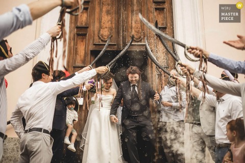 Newlyweds Exit Church Through Sword Gauntlet At Bergamo Lombardy Italy Pirates-Themed Wedding In Bergamo, Lombardy, Italy, newlyweds exit the church and walk through a gauntlet of swords held by friends, a nod to their wedding's "pirates" theme. The winning photograph highlights the photographer's skill at capturing unique, unscripted moments.