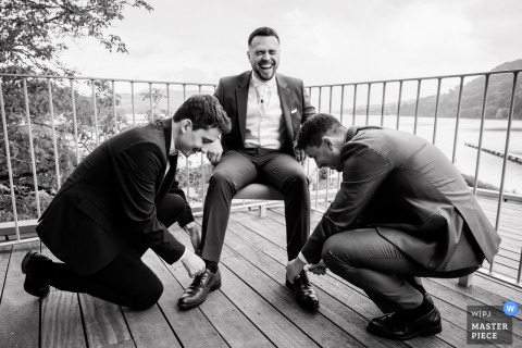 At a reception in Hérault, Occitanie, the photographer captures the groom erupting with laughter as his groomsmen kneel to tie his shoes, exemplifying timing and candid storytelling.