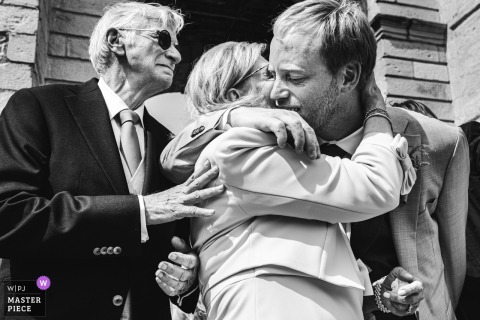 At Eglise d'Urrugne in Pyrénées Atlantiques, France, the photographer captures a moving low-angle black-and-white shot as the groom embraces his parents outside the church, expertly highlighting deep emotion and familial connection.