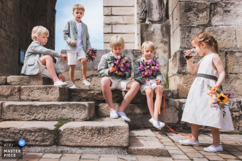 At Eglise d'Urrugne in Pyrénées Atlantiques, France, the photographer captures the young attendants waiting patiently on the church steps after the religious ceremony, highlighting composed innocence and skillful storytelling.