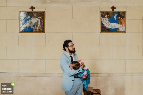 Father Tenderly Carries His Sleeping Child During A Traditional Religious Wedding Ceremony In Meuse France Inside a quiet church in Meuse, a father tenderly carries his sleeping child while standing against a stone wall with two paintings during a traditional and reverent religious wedding ceremony in the Grand Est.