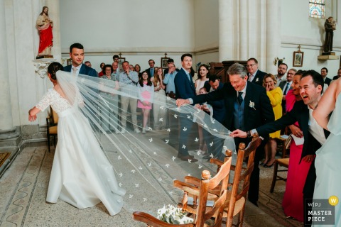 In Herault, at the church, the photographer captures the bride with her veil caught in a chair, skillfully preserving a lighthearted, candid moment and showcasing a keen eye for charming, unscripted wedding day details.