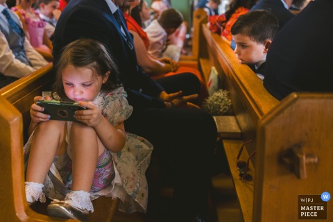 At a County Wicklow ceremony in Leinster, the photographer captures a girl seated on a church pew, engrossed in her phone, skillfully highlighting candid modern moments amid traditional wedding surroundings.